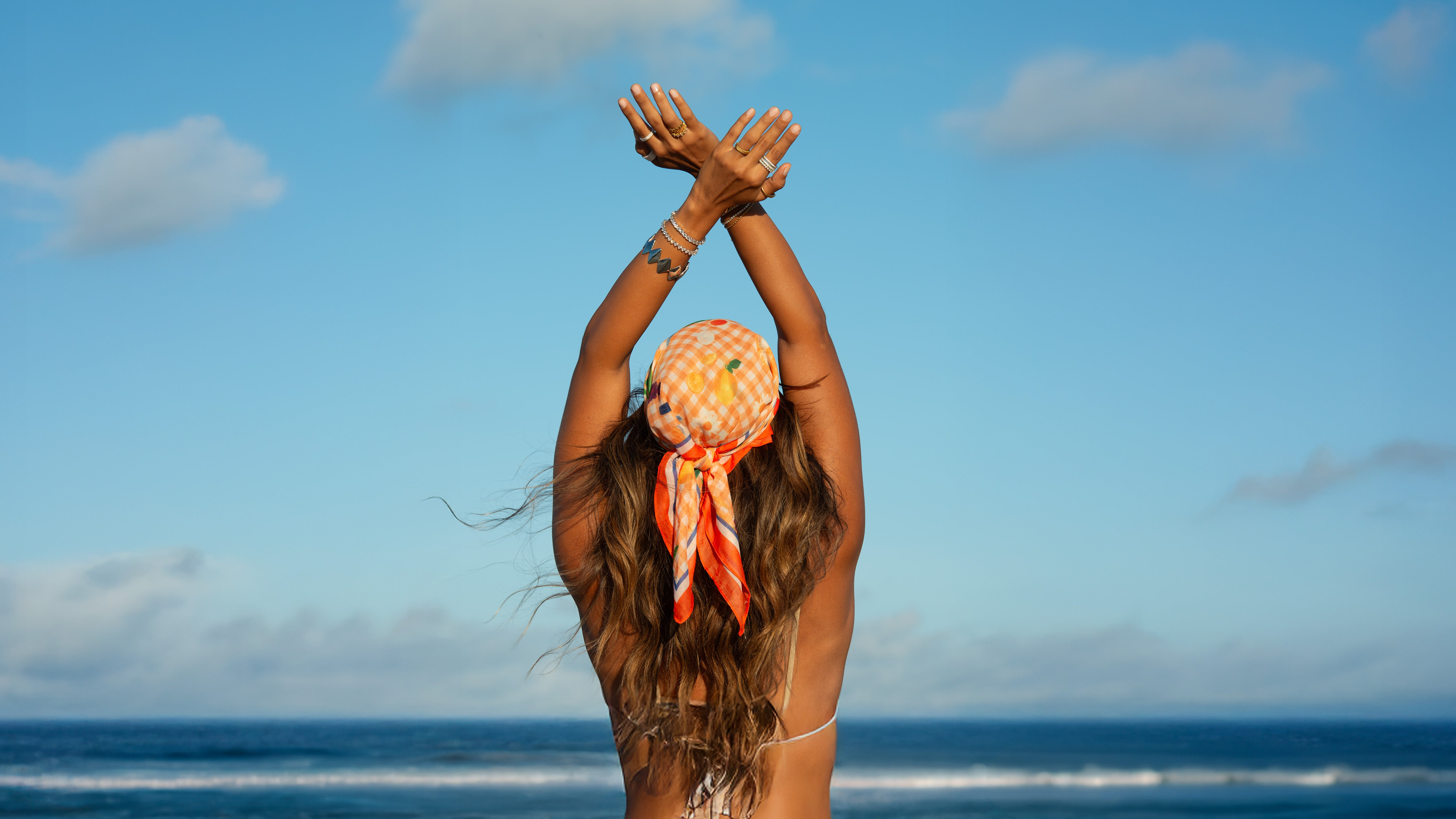 A woman seen from behind on a beach, wearing a patterned orange and white headscarf and a white bikini top, with her arms raised above her head against a blue sky and ocean. She wears Divé waterproof jewellery in silver, tennis bracelets, rings with zirconia stones, diamond-like sparkle and iconic geometric Edgy bracelet designed by Rebeca Sá